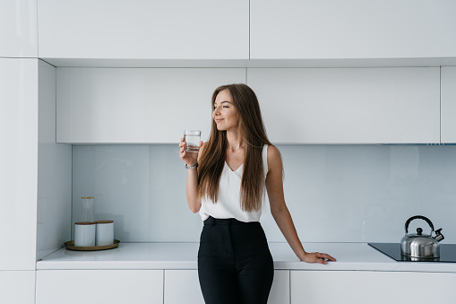 Satisfied fit businesswoman in elegant clothes standing at kitchen holds glass of water looks aside smiles. Attractive Italian female satisfied by healthy lifestyle. Successful entrepreneur dreaming.