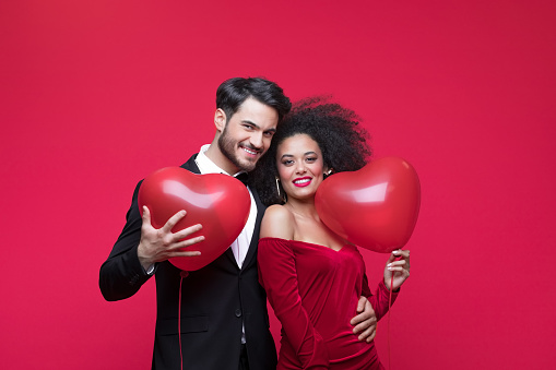 Young couple wearing elegant, woman in red dress, man in suit, holding heart shaped balloons embracing and smiling at camera. Studio shot, red background.