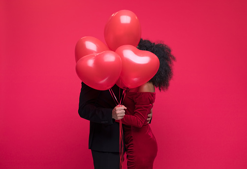 Young couple wearing elegant covering with bunch of heart shaped balloons. Studio shot, red background.