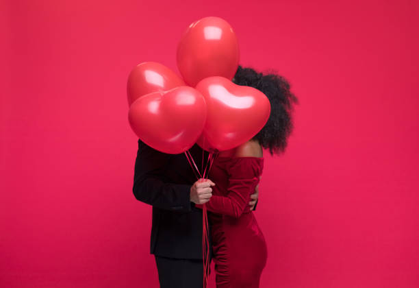 Beautiful couple celebrating Valentine's Day Young couple wearing elegant covering with bunch of heart shaped balloons. Studio shot, red background. be my valentine stock pictures, royalty-free photos & images