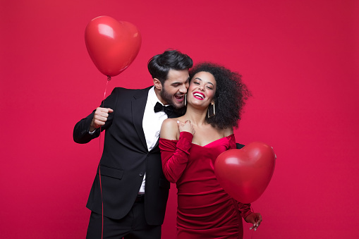 Young couple wearing elegant, woman in red dress, man in suit, holding heart shaped balloons embracing and smiling at camera. Studio shot, red background.