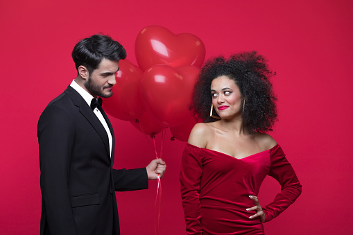 Young couple wearing elegant, woman in red dress, man in black suit, holding heart shaped balloons, looking at each other and smiling. Studio shot, red background.