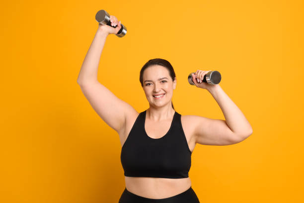istock Happy overweight woman doing exercise with dumbbells on orange background 1454154308