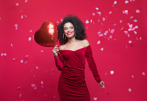 Young woman wearing red elegant dress, holding heart shaped balloon, looking away and smiling, standing among confetti. Studio shot, red background.