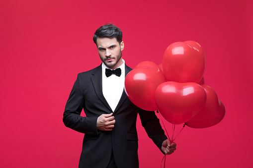 Young man wearing elegant black suit, holding heart shaped balloons and looking at camera. Studio shot, red background.