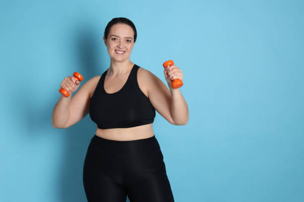 istock Happy overweight woman doing exercise with dumbbells on light blue background. Space for text 1454155755