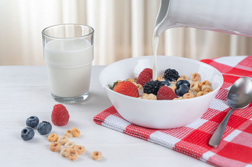 Pouring fresh milk into a bowl with muesli and berries, on a red checkered napkin on a white table