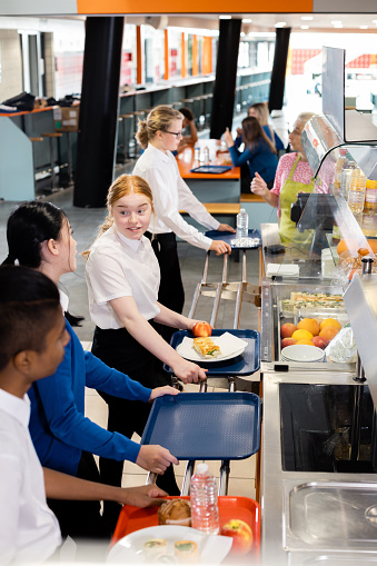 A side view of a group of high school children who are waiting in line at the food bar to get their lunch in a school in the North East of England. At the food bar there is healthy snacks and drinks. They are chatting to each other as they wait in line.