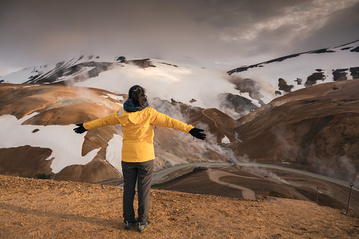 Cheerful asian woman in yellow jacket standing on geothermal area and volcanic mountain on summer in highlands at Kerlingarfjoll, Iceland
