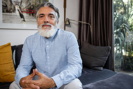 A serious senior man with gray hair at home, sitting on the couch and looking at camera