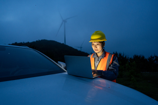 A female worker uses a computer to work in front of the car at the wind farm at night