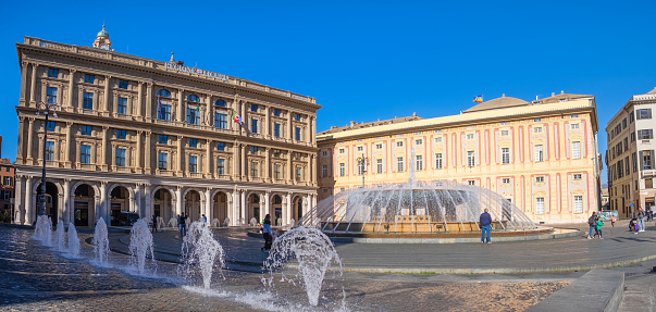 Tourists visiting Piazza De Ferrari in Genoa, the main city square characterized by a large circular fountain and many historic buildings, such as the Palazzo Ducale and the Palazzo della Regione (2 shots stitched)