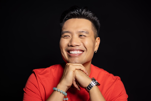 Close-up portrait of smiling asian man on black background. Positive man in red t-shirt with hands clasped looking away in studio.