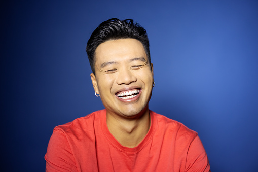 Close-up portrait of smiling young Vietnamese man on blue background. Happy male in red t-shirt smiling in studio.