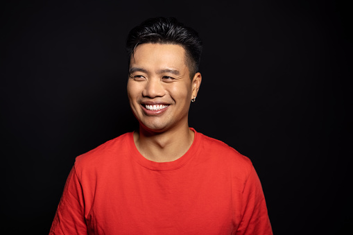 Portrait of confident young asian man looking away. Man in red t-shirt smiling on black background.