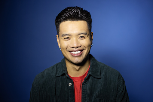 Close-up portrait of a happy young Asian man on blue background. Confident male in casuals looking at camera and smiling in the studio.