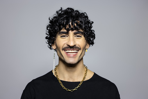 Studio portrait of a gender fluid person smiling on grey background. Close-up of a mid-adult wearing earrings looking at camera in studio.