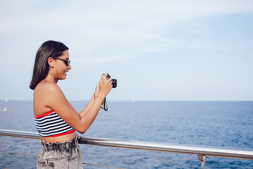 Side view of cheerful ethnic female tourist in sunglasses with photo camera standing on quay and leaning on metal railing while taking pictures of sea during summer vacation