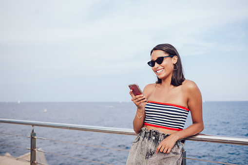 Smiling traveling female in sunglasses and summer outfit reading messages while using smartphone and leaning on metal railing at seafront