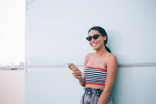 Modern happy ethnic brunette in stylish sunglasses and top holding smartphone and looking brightly at camera standing against street wall