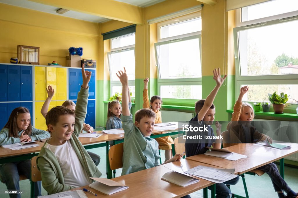 Children learning in the elementary school Boys and girls sitting at the desks in the classroom, raising hands and answering. Children in the elementary school. Classroom Stock Photo Children learning in the elementary school Boys and girls sitting at the desks in the classroom, raising hands and answering. Children in the elementary school. Classroom Stock Photo