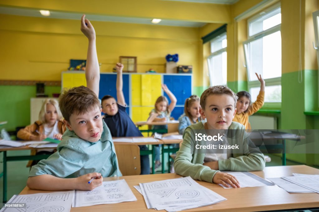 Children learning in the elementary school Boys and girls sitting at the desks in the classroom, raising hands and answering. Children in the elementary school. Classroom Stock Photo Children learning in the elementary school Boys and girls sitting at the desks in the classroom, raising hands and answering. Children in the elementary school. Classroom Stock Photo