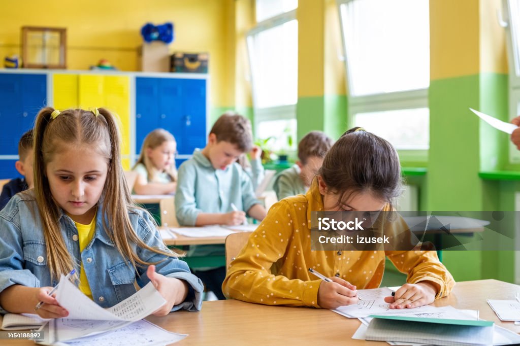 Children learning in the elementary school Cute girls sitting at the desk in the classroom and learning. School children in the background. Classroom Stock Photo Children learning in the elementary school Cute girls sitting at the desk in the classroom and learning. School children in the background. Classroom Stock Photo