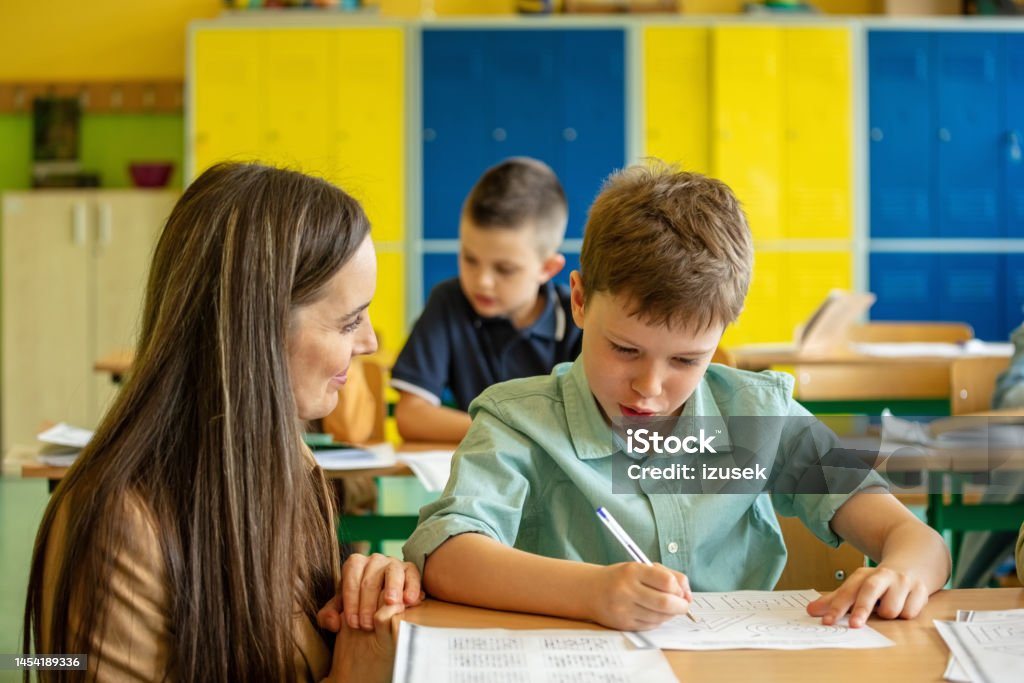 Female teacher helping boy during lesson School boy sitting at the desk in the classroom and learning with the female teacher. Children in the elementary school. 8-9 Years Stock Photo Female teacher helping boy during lesson School boy sitting at the desk in the classroom and learning with the female teacher. Children in the elementary school. 8-9 Years Stock Photo