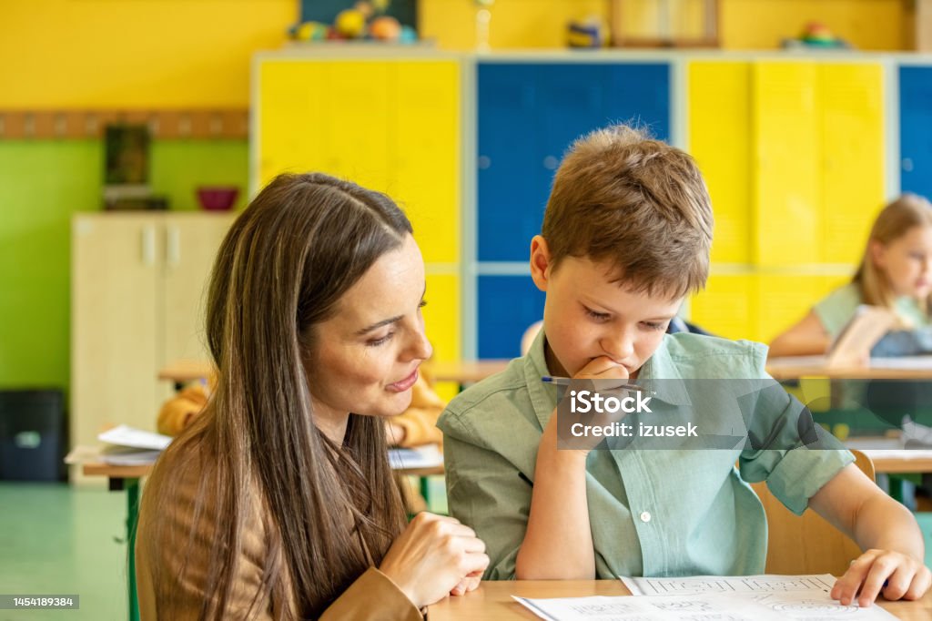 Female teacher helping boy during lesson School boy sitting at the desk in the classroom and learning with the female teacher. Children in the elementary school. 8-9 Years Stock Photo Female teacher helping boy during lesson School boy sitting at the desk in the classroom and learning with the female teacher. Children in the elementary school. 8-9 Years Stock Photo