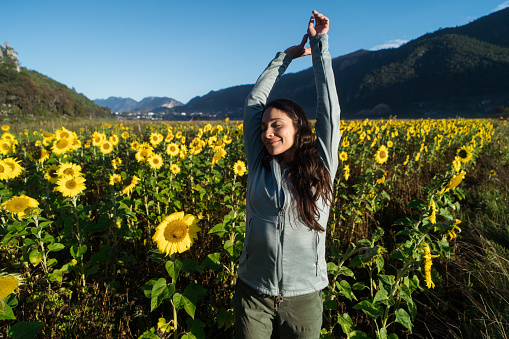 A young latin woman standing in front of a sunflower field with her eyes closed and arms stretched out.