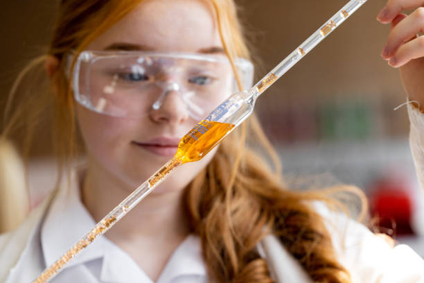 Science Experiment in School Close-up of a female teenager holding up a pipette filled with an orange substance. She is wearing a lab coat and protective eyewear. chemistry class stock pictures, royalty-free photos & images