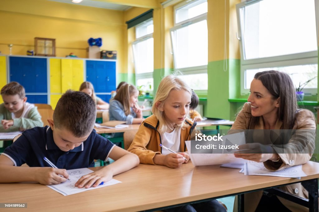 Female teacher helping boy during lesson School boys sitting at the desk in the classroom and learning with the female teacher. Children in the elementary school. Teacher Stock Photo Female teacher helping boy during lesson School boys sitting at the desk in the classroom and learning with the female teacher. Children in the elementary school. Teacher Stock Photo