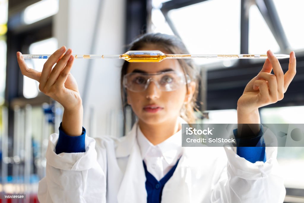 Concentrating in School Close-up of a female teenager holding up a pipette filled with an orange substance. She is wearing a lab coat and protective eyewear. Science Stock Photo