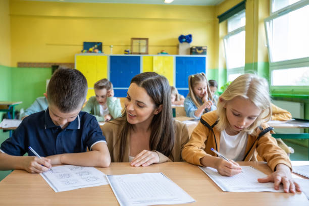 Female teacher helping boys during lesson School boys sitting at the desk in the classroom and learning with the female teacher. Children in the elementary school. schools expats stock pictures, royalty-free photos & images