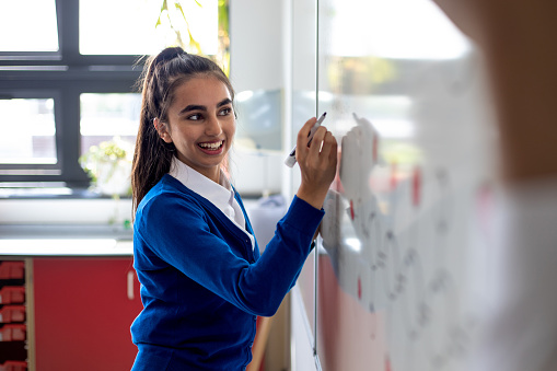 Waist-up shot of a female teenager writing on a whiteboard at her school with a smile on her face. She is wearing her school uniform.