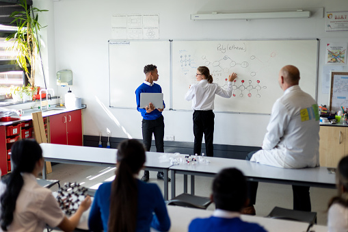 Wide shot of a science classroom with two male teenagers at the front of the class explaining something to the rest of the class. Everybody is listening and looking toward the board.