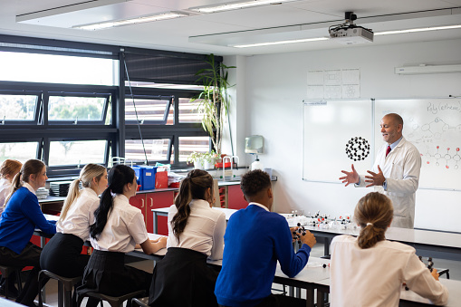 A wide shot of a teacher throwing up a molecular structure in the air whilst explaining it. The teacher is wearing a lab coat.