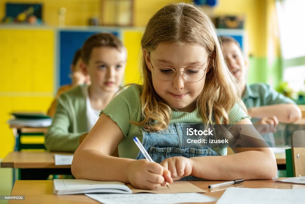 Schoolgirl sitting in the classroom Portrait of cute girl sitting at the desk in the classroom and writing. School children in the background. Classroom Stock Photo Schoolgirl sitting in the classroom Portrait of cute girl sitting at the desk in the classroom and writing. School children in the background. Classroom Stock Photo