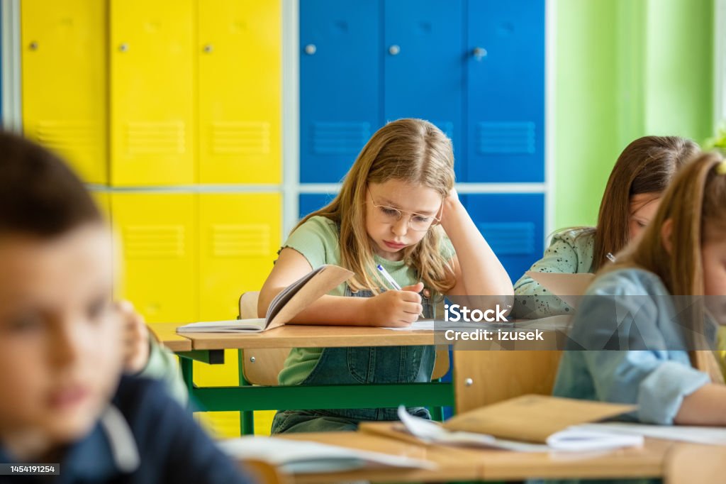 Schoolgirl sitting in the classroom Portrait of cute girl sitting at the desk in the classroom and writing. Children learning in the elementary school. Child Stock Photo Schoolgirl sitting in the classroom Portrait of cute girl sitting at the desk in the classroom and writing. Children learning in the elementary school. Child Stock Photo
