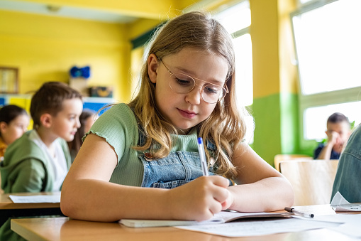 Portrait of cute girl sitting at the desk in the classroom and writing. School children in the background.