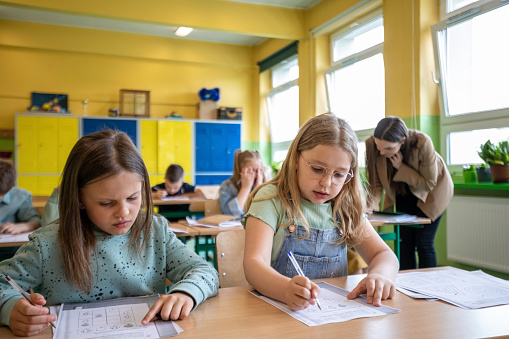 Girls sitting at the desk in the classroom and learning together. School children and female teacher in the background.