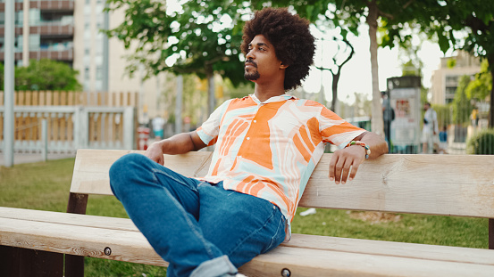 Close-up of young African American man wearing shirt sitting relaxed on park bench looking away. Life style concept.
