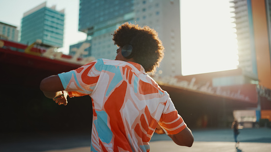 Close-up of cheerful young African American man wearing shirt listening to music in headphones and dancing on urban city background. Lifestyle concept