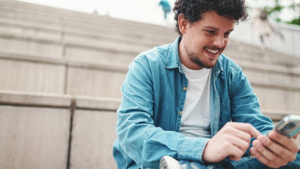 istock smiling young bearded man in denim shirt sitting on high steps and using mobile phone. Man playing on smartphone 1454193515