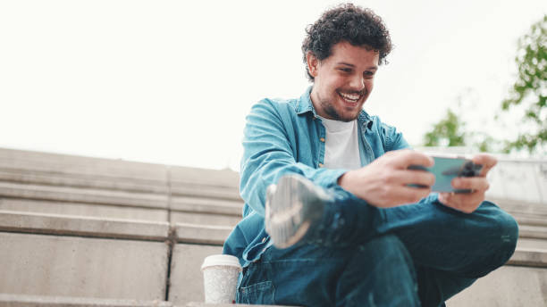 istock smiling young bearded man in denim shirt sitting on high steps and using mobile phone. Man playing on smartphone 1454193516