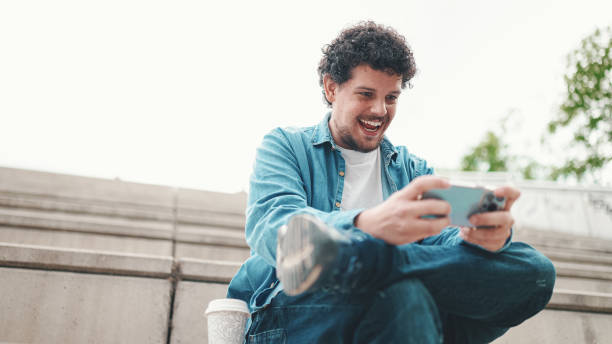 istock smiling young bearded man in denim shirt sitting on high steps and using mobile phone. Man playing on smartphone 1454193518