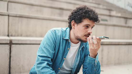 young bearded man in denim shirt sitting on high steps and using mobile phone. man sends voice message to smartphone