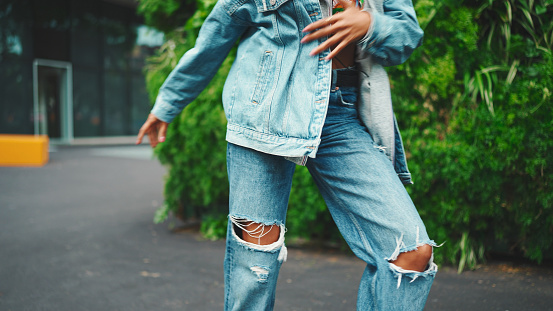 Closeup, smiling African girl wearing denim jacket, in crop top with national pattern walks in city park listening to music on headphones and dancing on green trees background.
