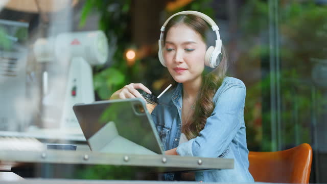 smiling cheerful relax asian female woman enjoy watch streaming movie music from tablet and wearing headphone sit next to cafe window shopfront morning weekend routine shoot through window coffeeshop