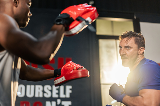 Caucasian man wear boxing gloves punching ahead with African trainer. Attractive young body builder boxing male working out with help of African American coach for health care in gym or fitness club.
